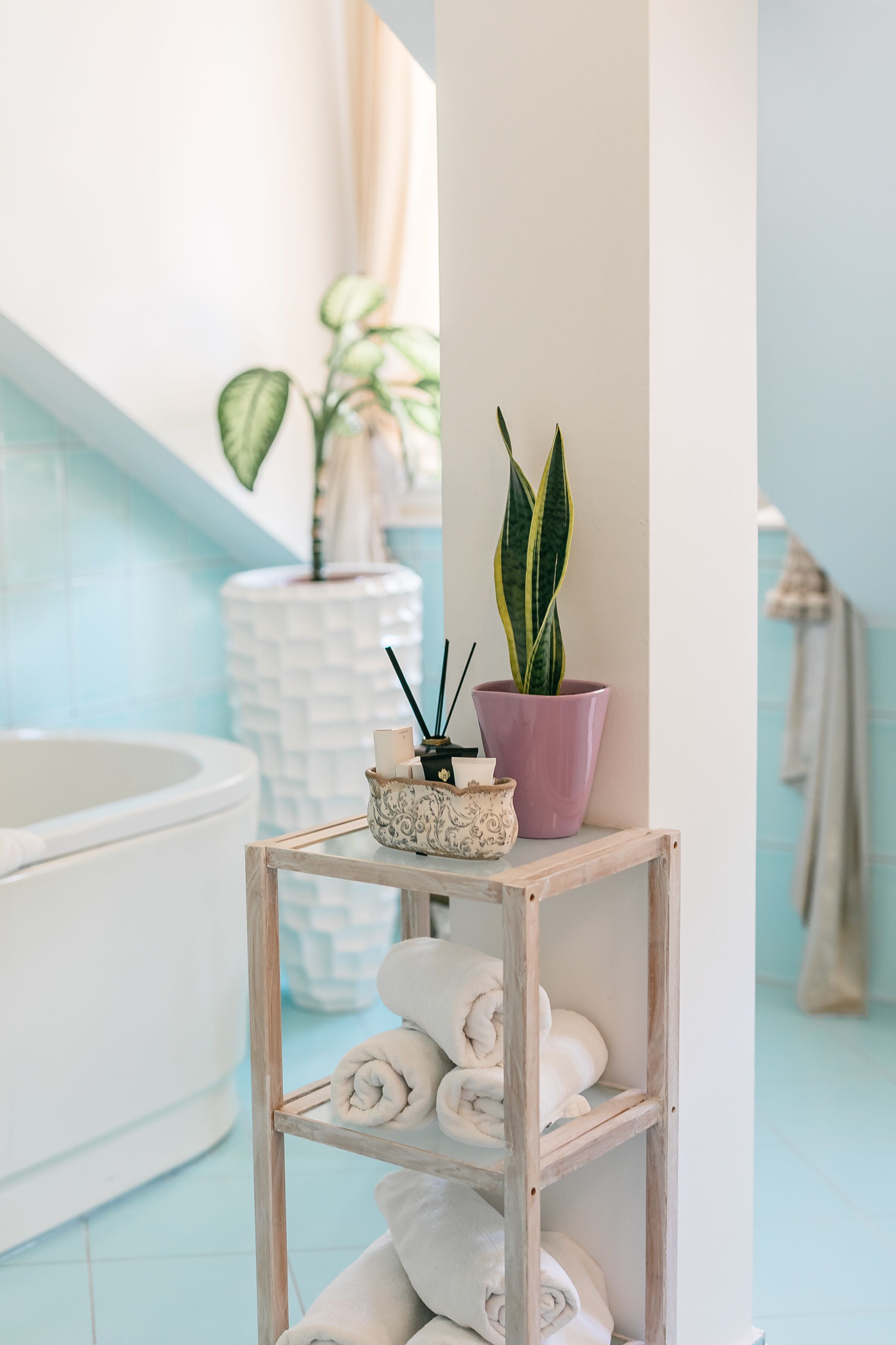 Un baño con una bañera blanca, un estante de madera con toallas enrolladas, una planta en maceta y un florero blanco con velas.