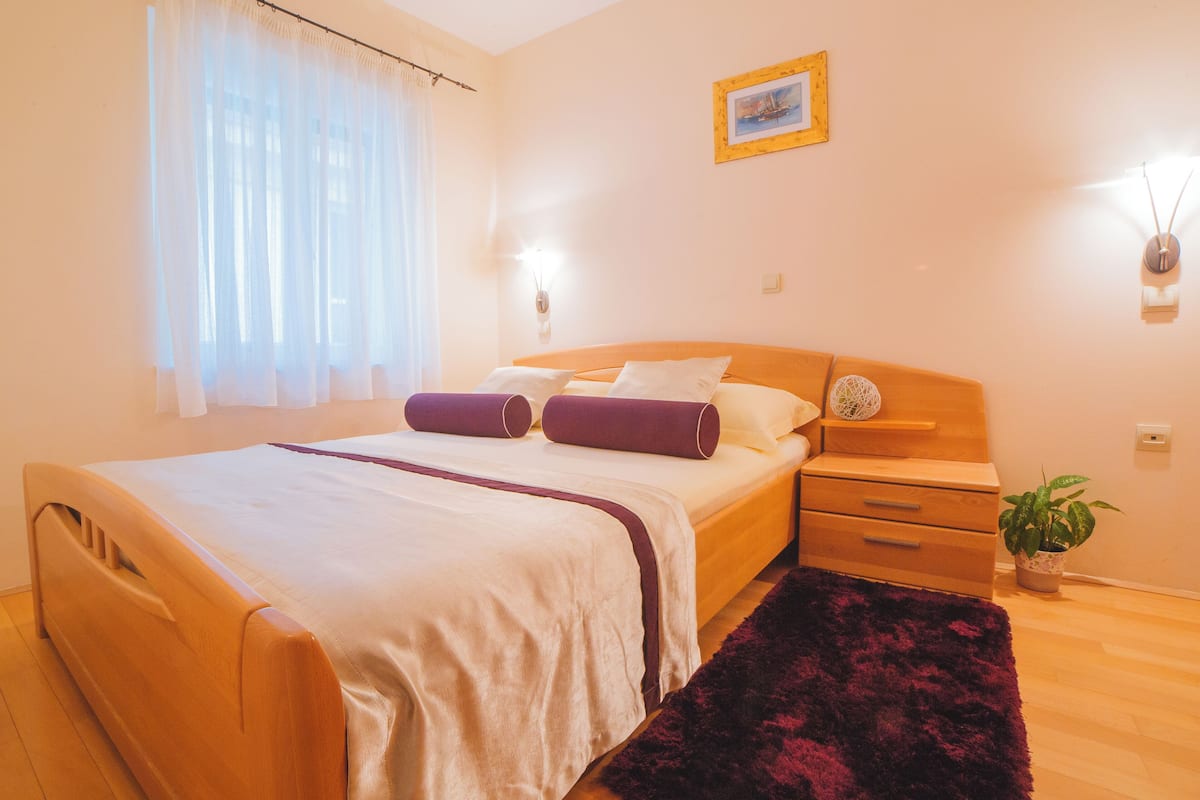 A neatly arranged bedroom with a bed, bedside tables, a nightstand, a potted plant, and a window with sheer curtains.