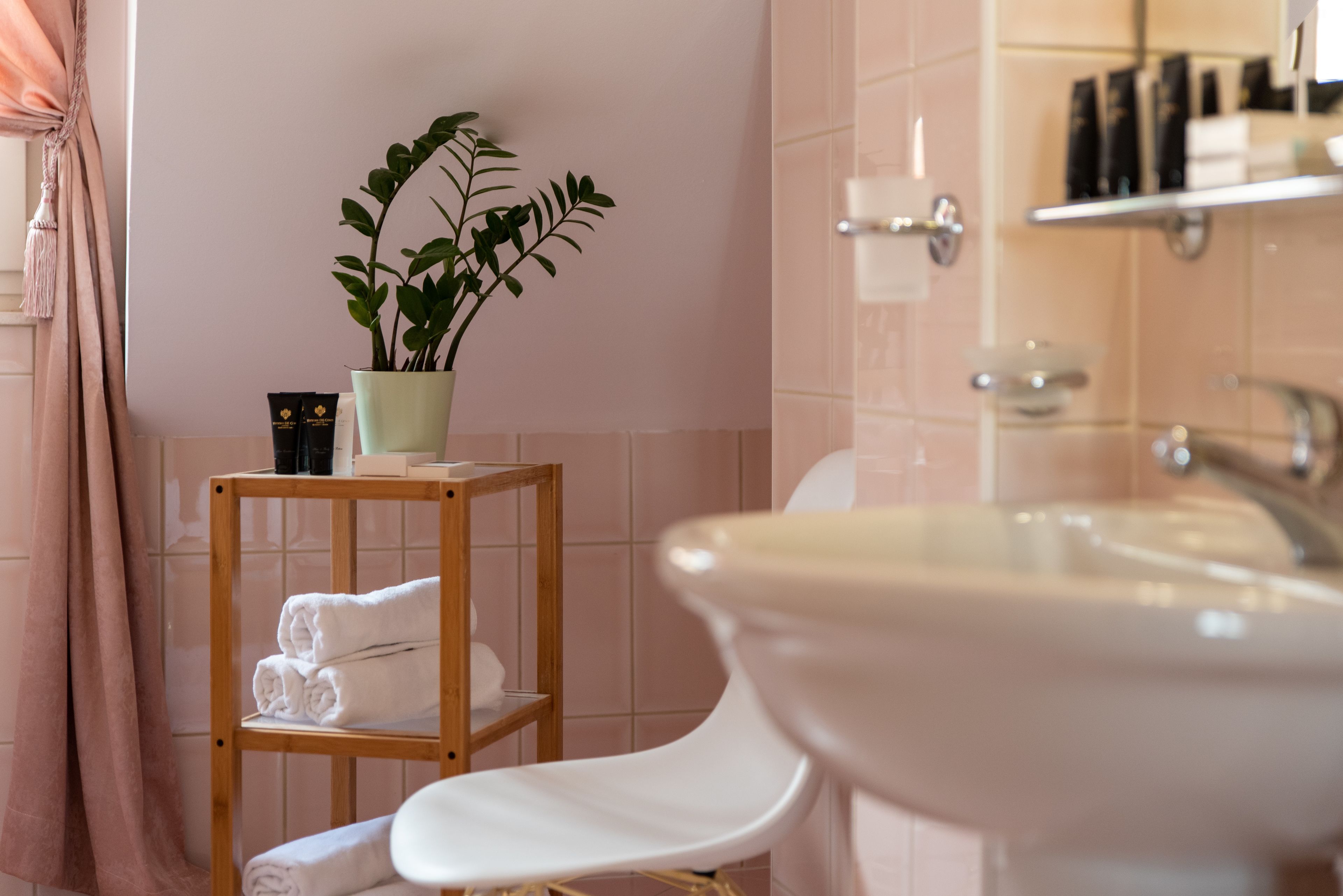 Une salle de bain avec une étagère en bois où sont posés des serviettes et des articles de toilette, une plante en pot et un grand lavabo.