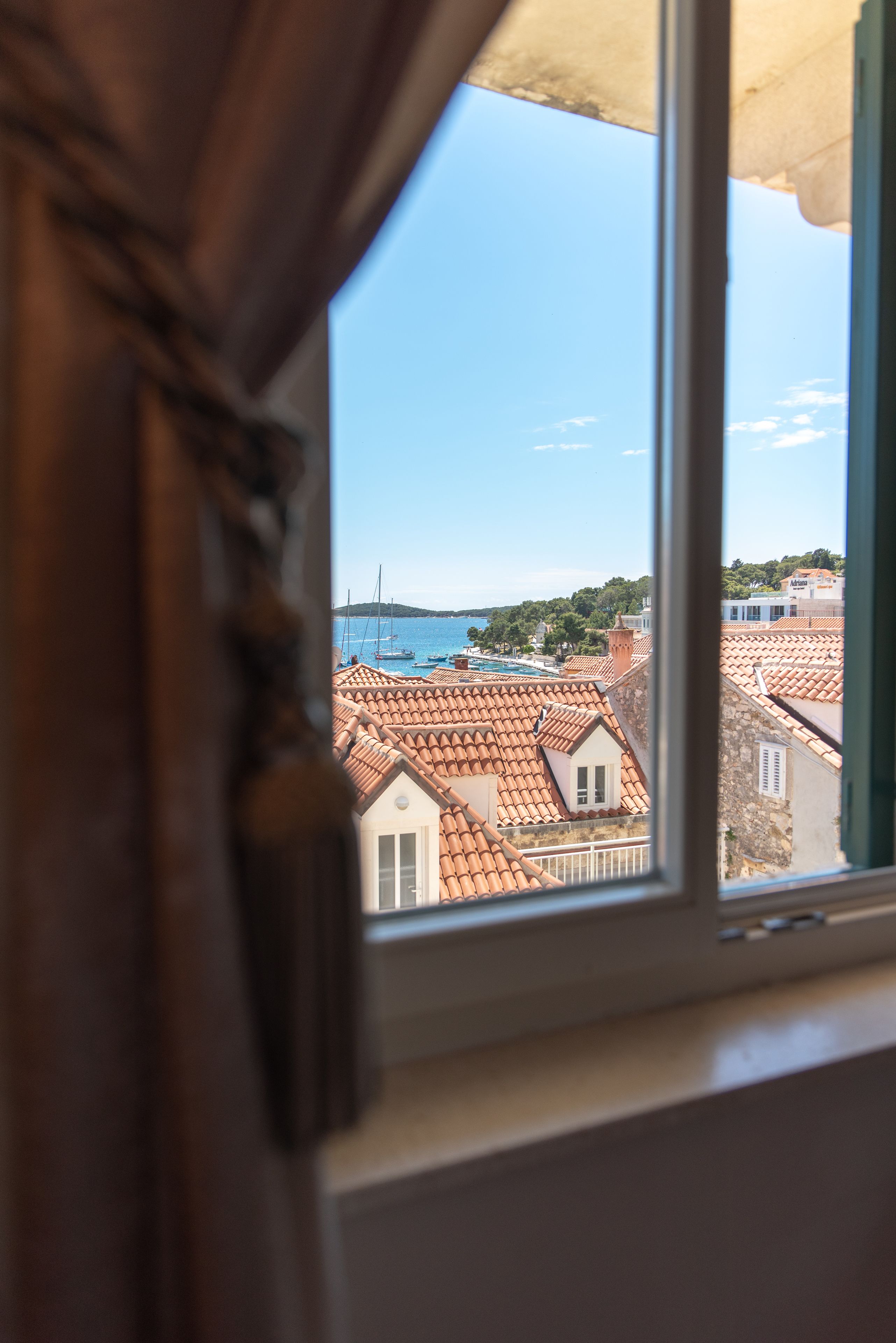 A view through a window with a curtain, revealing a coastal town with terracotta-roofed buildings and a clear blue sky.