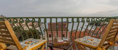 A balcony with two wooden chairs and a small table, overlooking a coastal town with red-roofed buildings and boats in the water.