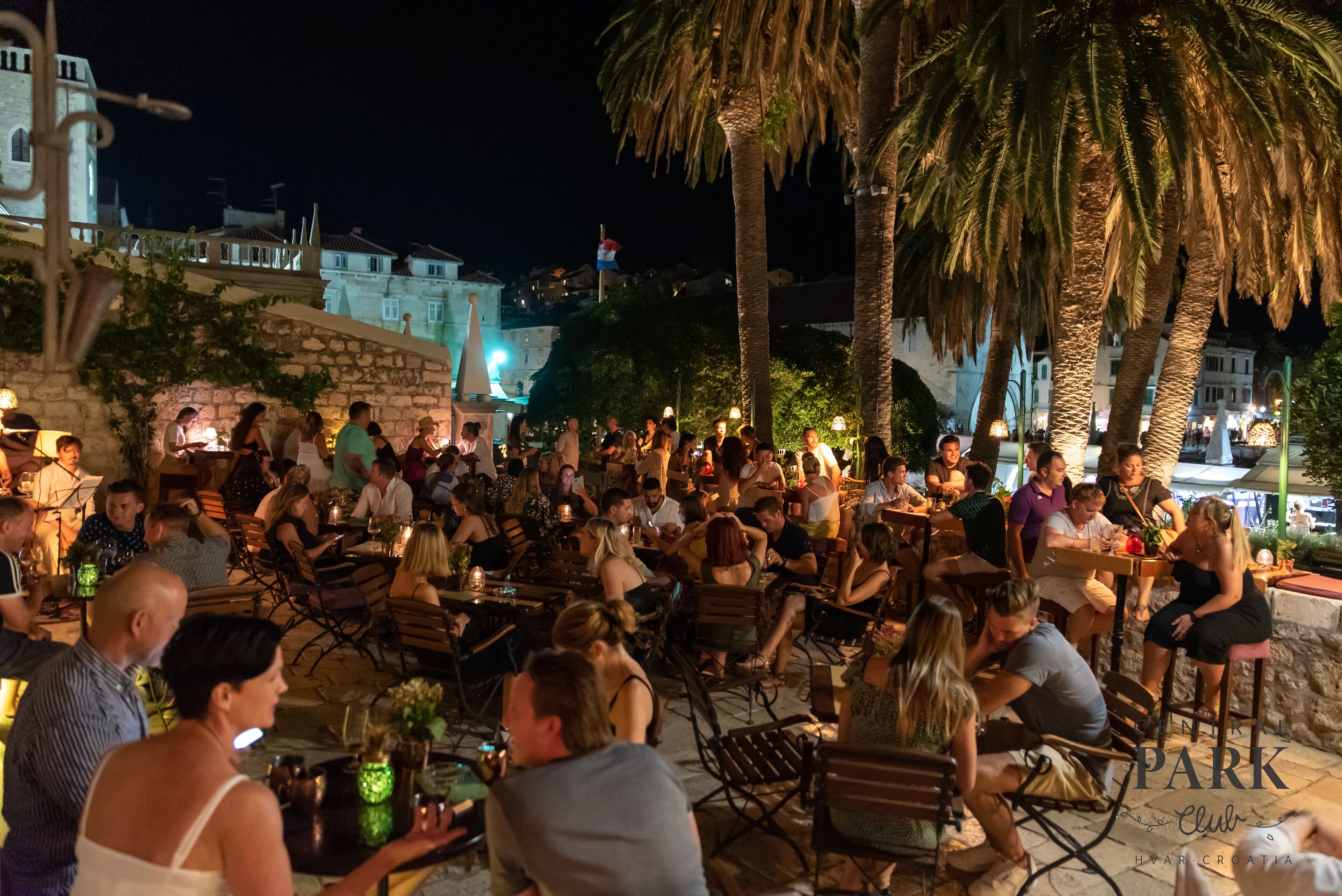 Een terras met stenen muren en palmbomen, waar een groep mensen aan tafels zitten.