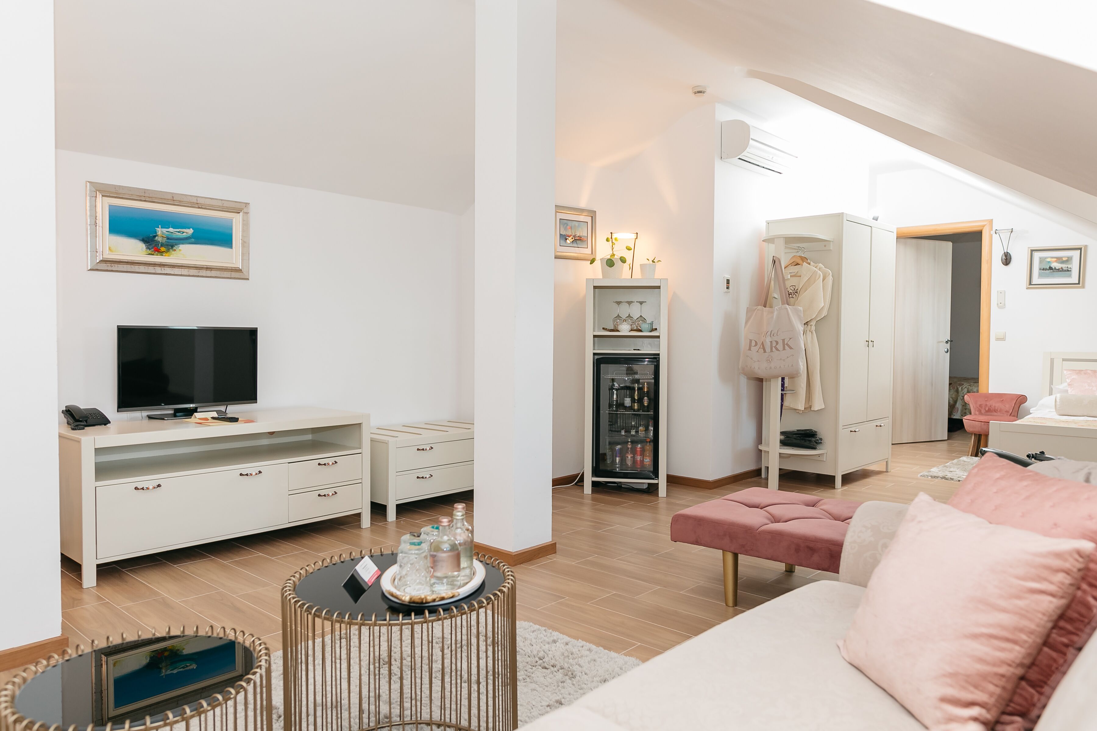 A modern living room with a flat-screen TV, white furniture, a wicker coffee table, and a pink ottoman.
