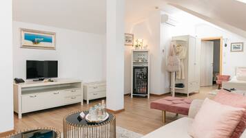 A modern living room with a flat-screen TV, white furniture, a wicker coffee table, and a pink ottoman.