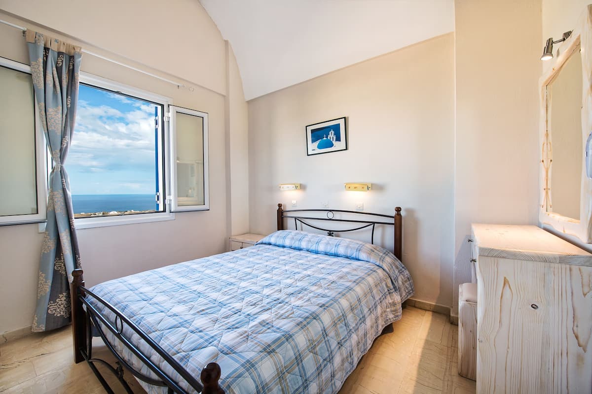 A bedroom with a bed, a window with curtains, a bedside table, a wall-mounted light, and a small cabinet.