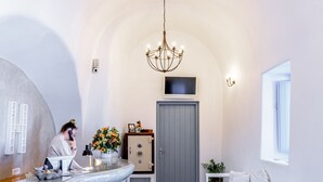 A modern interior with a curved reception desk, a white sofa, a chandelier, and a grey door. - Anatoli Hotel and Spa (Santorini)