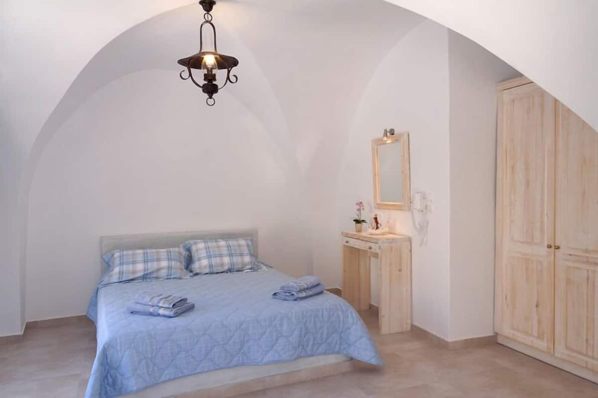 A bedroom with a bed, a wooden wardrobe, a bedside table with a mirror, and a hanging light fixture.