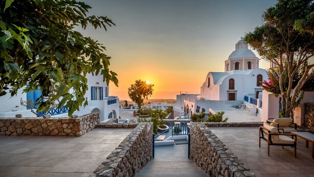 A stone staircase leading to a white building with a dome, surrounded by greenery and a sunset in the background. - Anatoli Hotel and Spa (Santorini)