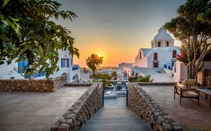 A stone staircase leading to a white building with a dome, surrounded by greenery and a sunset in the background. - Anatoli Hotel and Spa (Santorini)