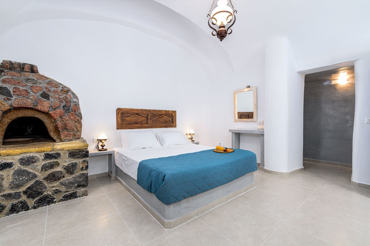 A bedroom with a stone fireplace, a bed with a blue blanket, a nightstand with a lamp, and a mirror on the wall.