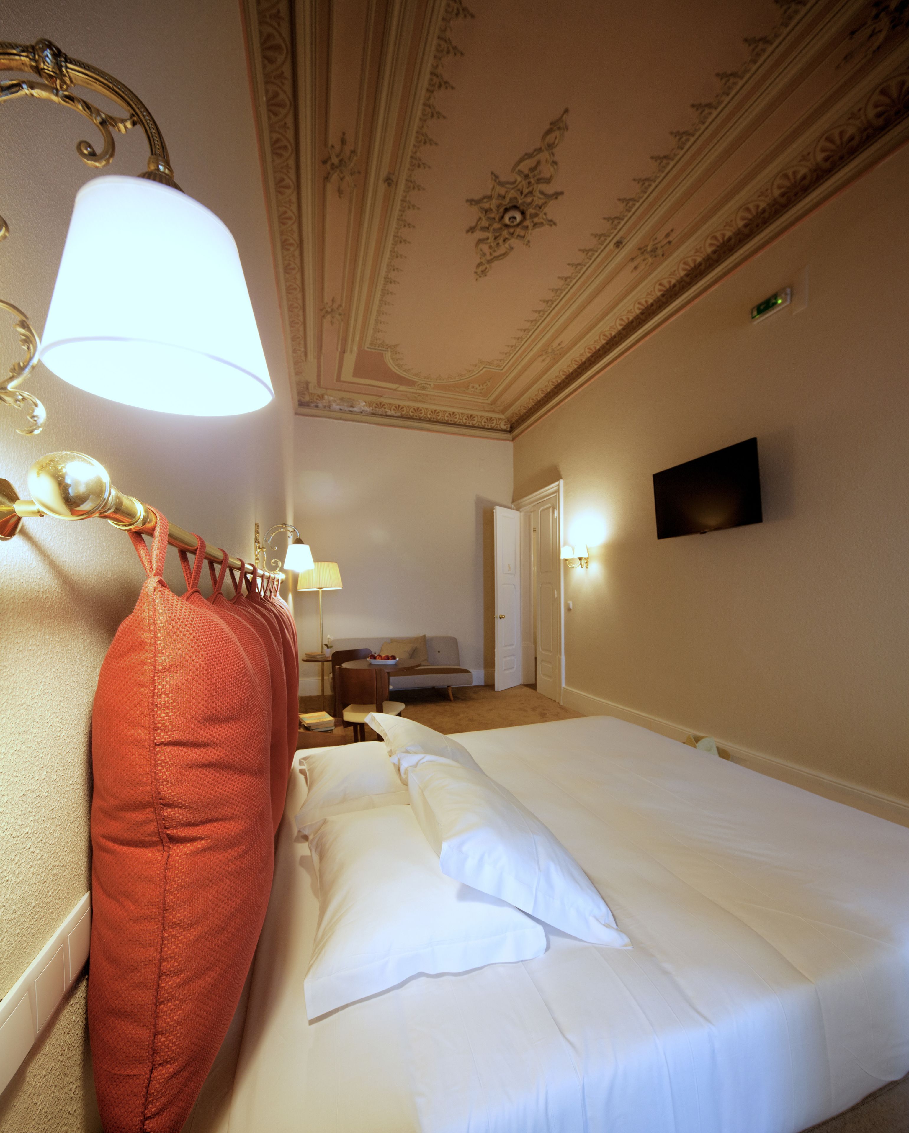 A hotel room with a bed, a red pillow, a lamp, a television, and a door.