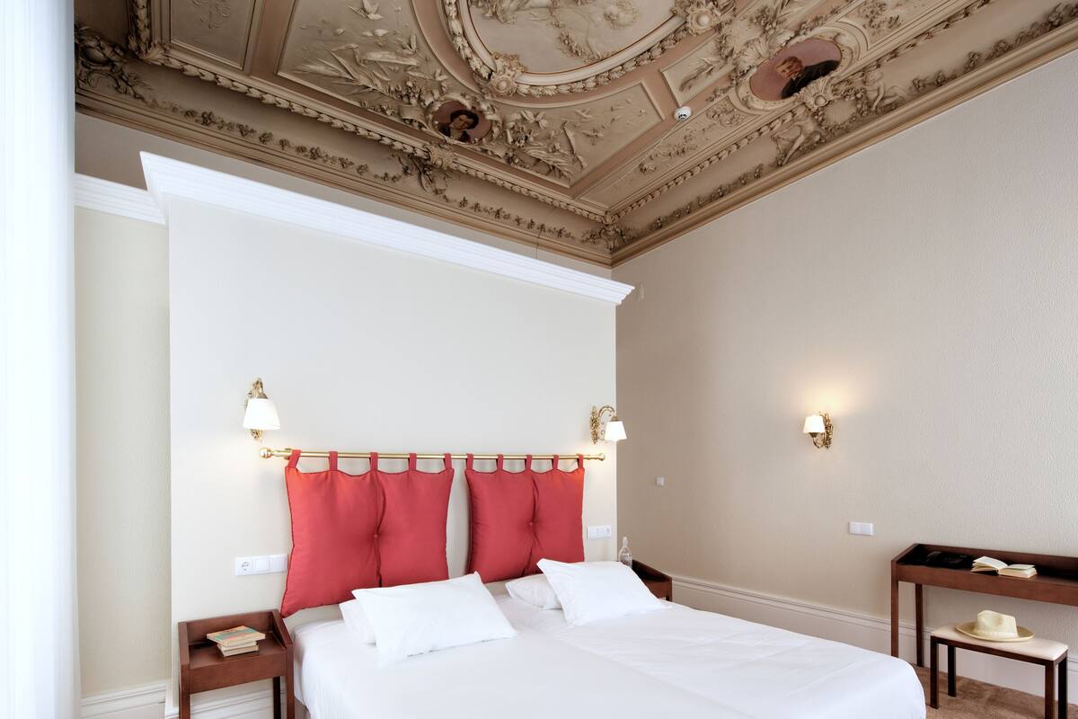 A hotel room with a large bed, red pillows, a decorative ceiling, and a small table with a hat.