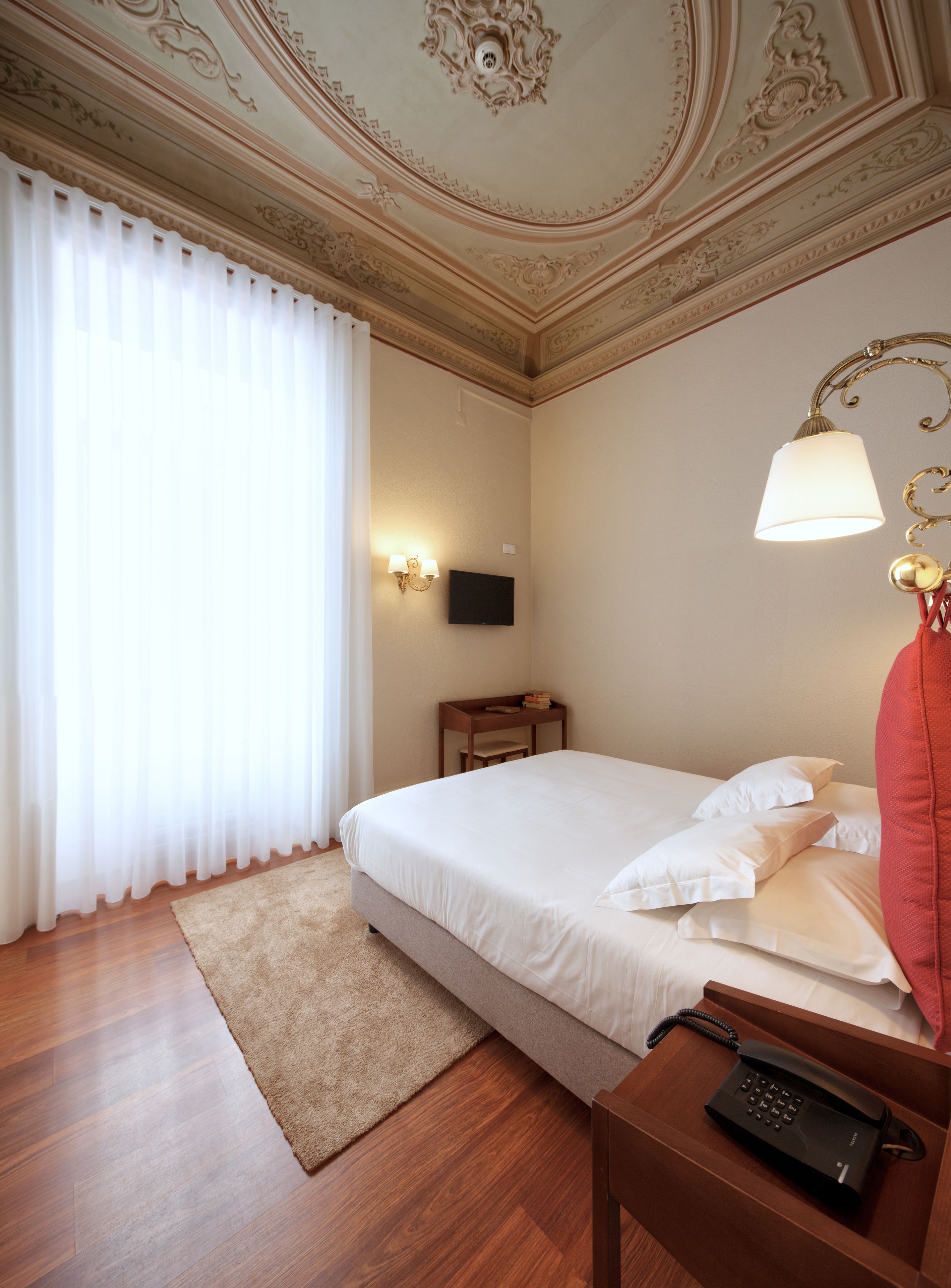 Habitación de hotel con cama, mesita de noche con lámpara, una pequeña mesa con libros y un televisor de pantalla plana montado en la pared.