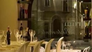 Un restaurant avec des chaises et des tables blanches, une vue sur un immeuble à travers une grande fenêtre, et des étagères remplies de bouteilles et de verres.