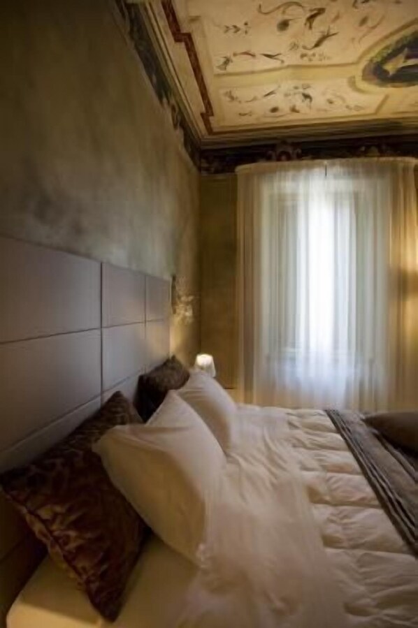 Una habitación de hotel con una cama grande, cabecera, mesita de noche y ventana con cortinas.