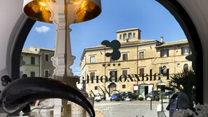 Une lampe avec un abat-jour blanc et un pied doré, qui reflète, à travers une fenêtre, la façade d’un immeuble sur lequel on peut lire l’inscription « Hôtel Blokxhoog ».