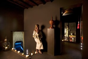 Une personne vêtue d’une robe se tient dans une pièce faiblement éclairée, où l’on aperçoit une cheminée, des bougies et un grand miroir qui reflète l’image d’une autre personne portant une robe.