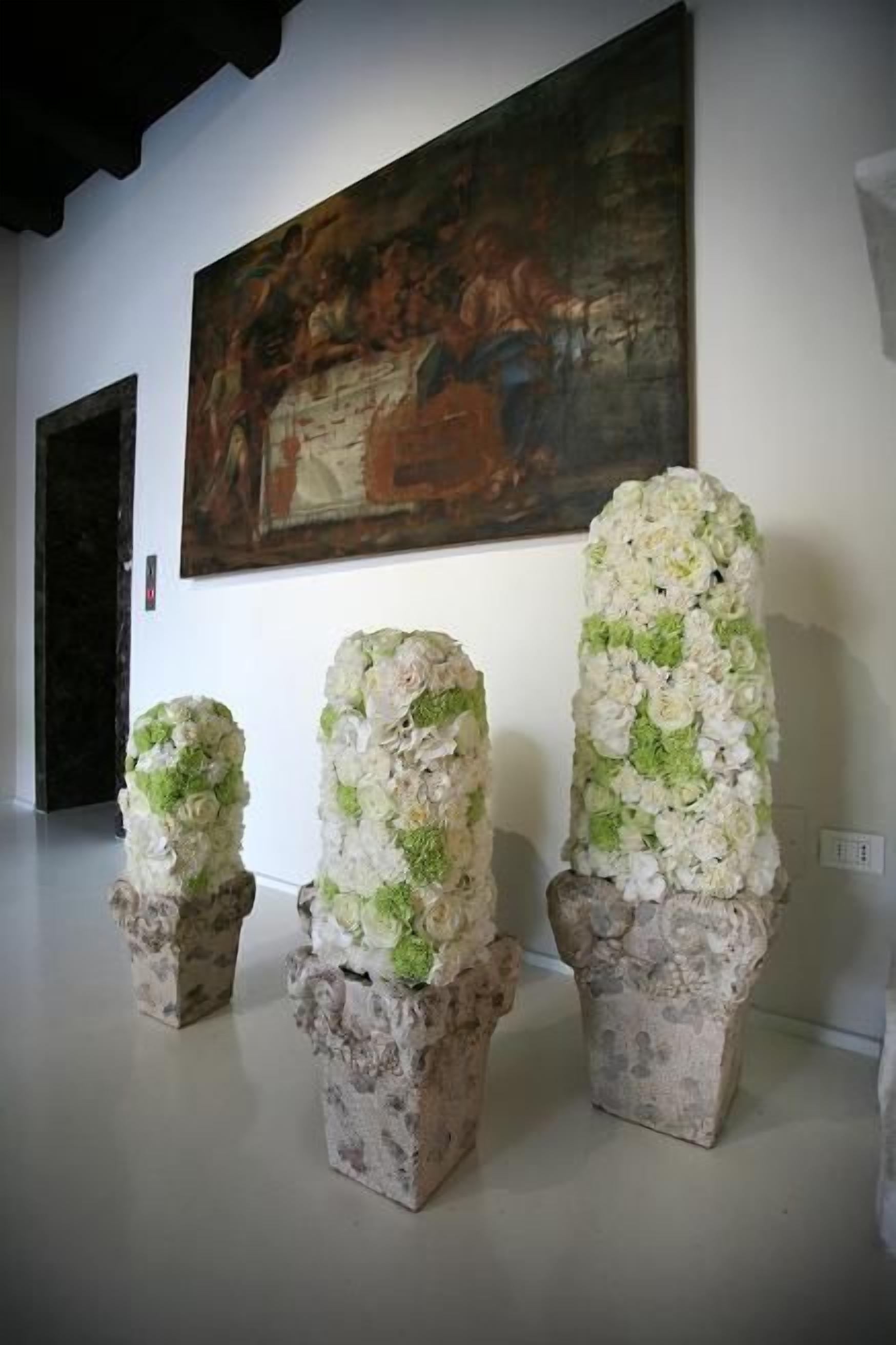 Tres arreglos florales sobre pedestales en una galería, con un cuadro en la pared.