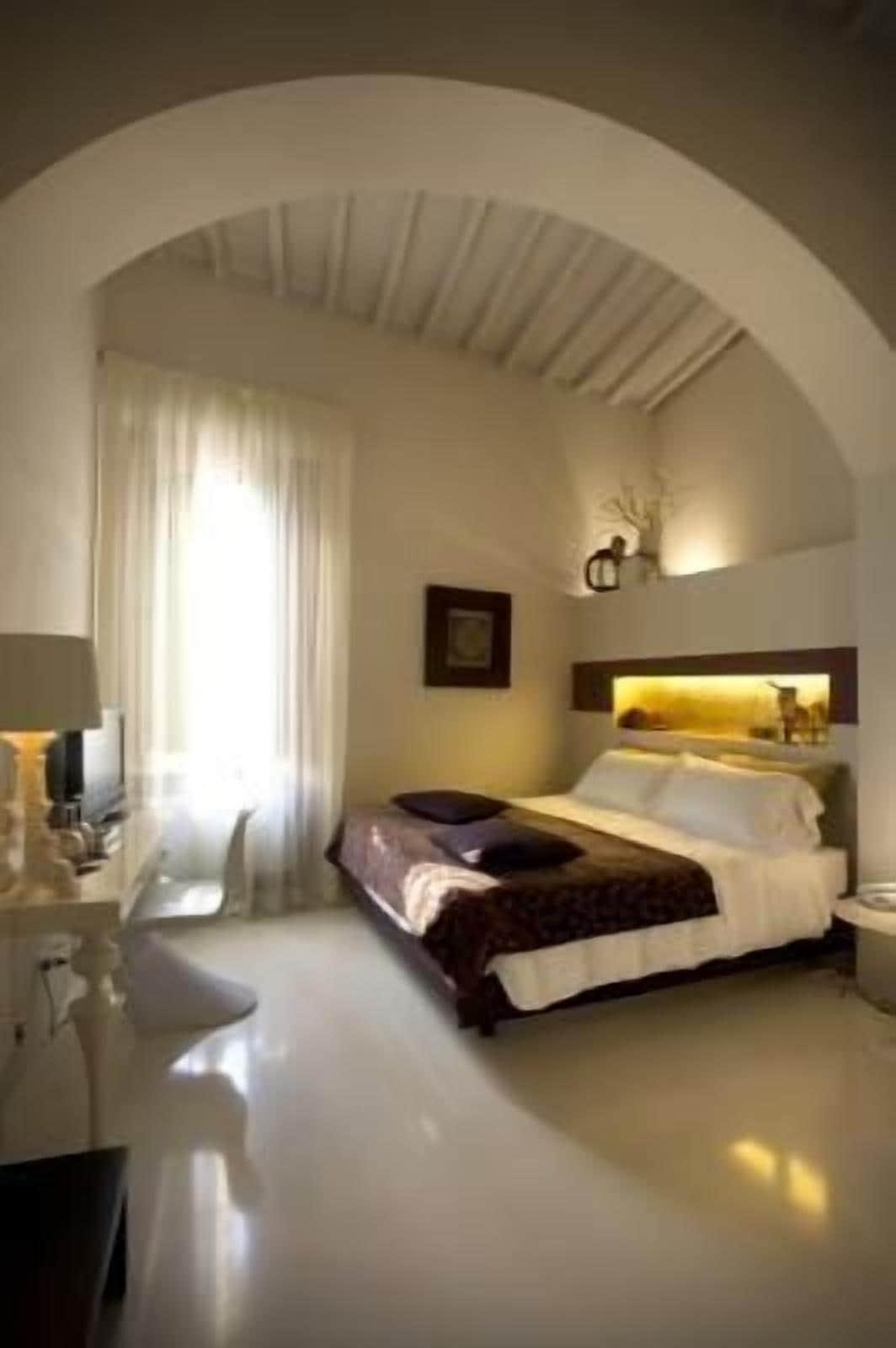 Ein Hotelzimmer mit Bett, Schreibtisch und Stuhl. Ein Fenster mit Vorhängen, ein Bild an der Wand und eine Lampe auf dem Schreibtisch.