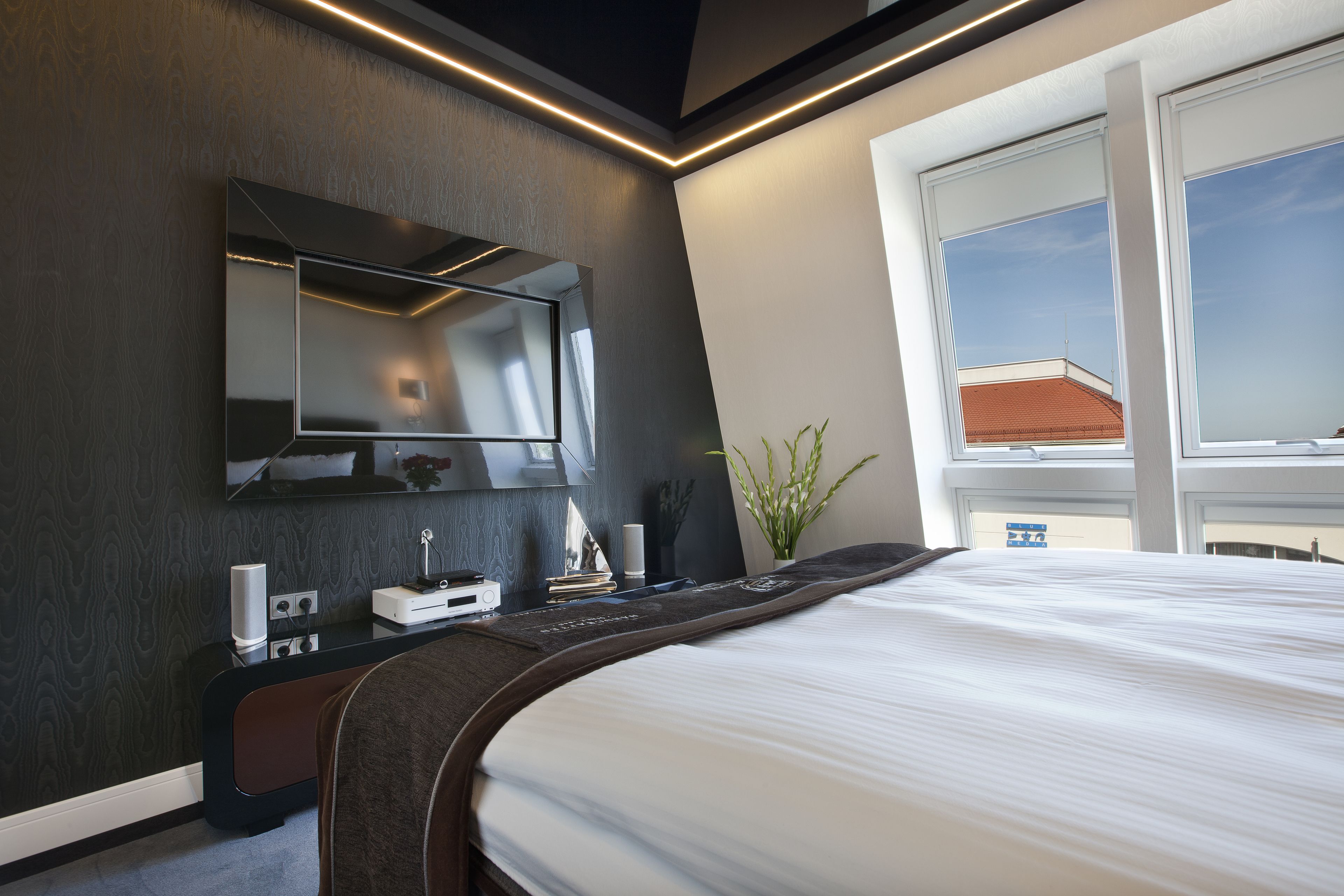 Una habitación de hotel moderna con una cama grande, un televisor de pantalla plana en la pared, una mesita de noche con lámpara y una ventana con vistas al exterior.