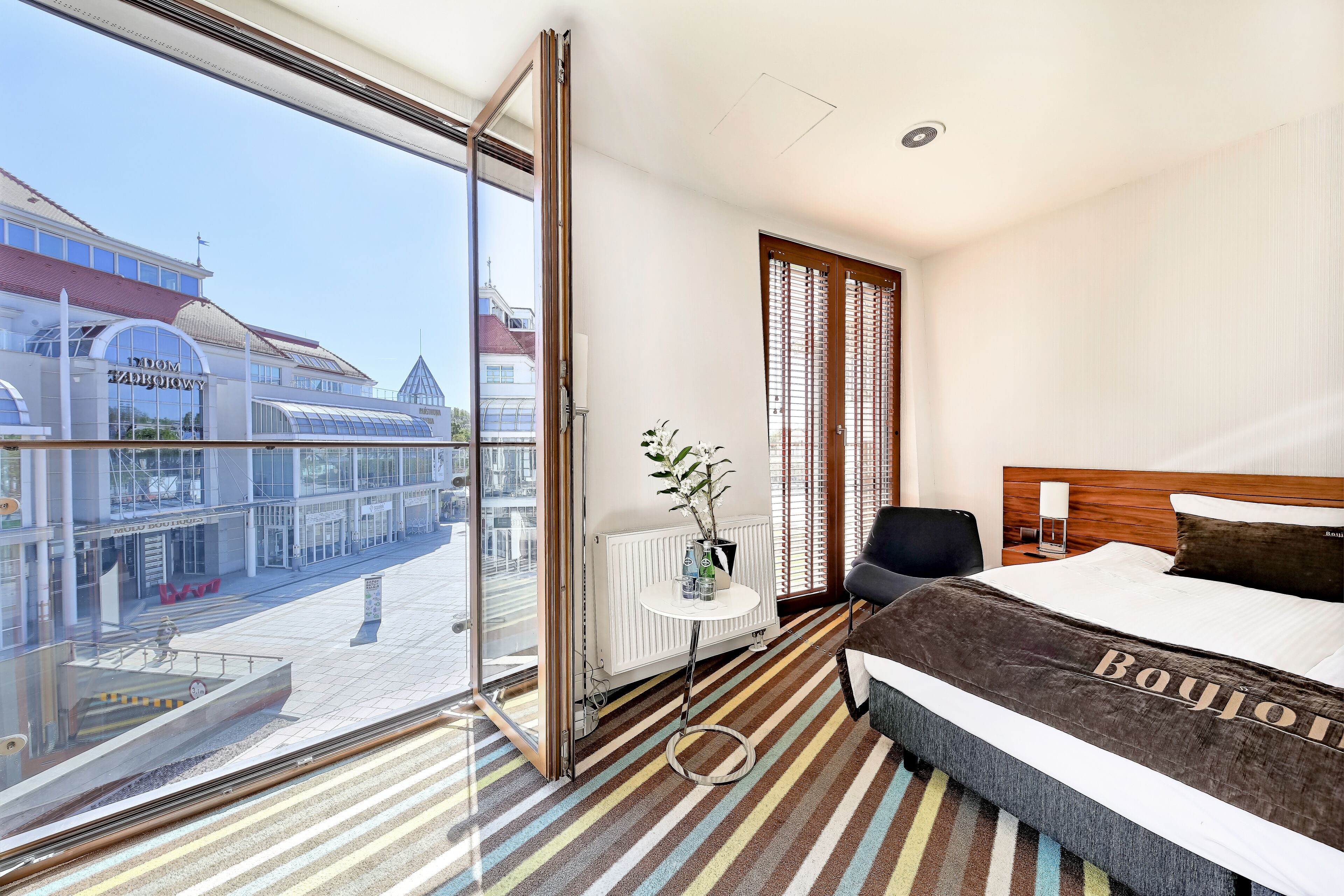 A hotel room with a large window, a bed with a 'Bayon' bedspread, a radiator, a small table with a plant, and a view of a building with a sign reading 'DOM TAROTOWY'.