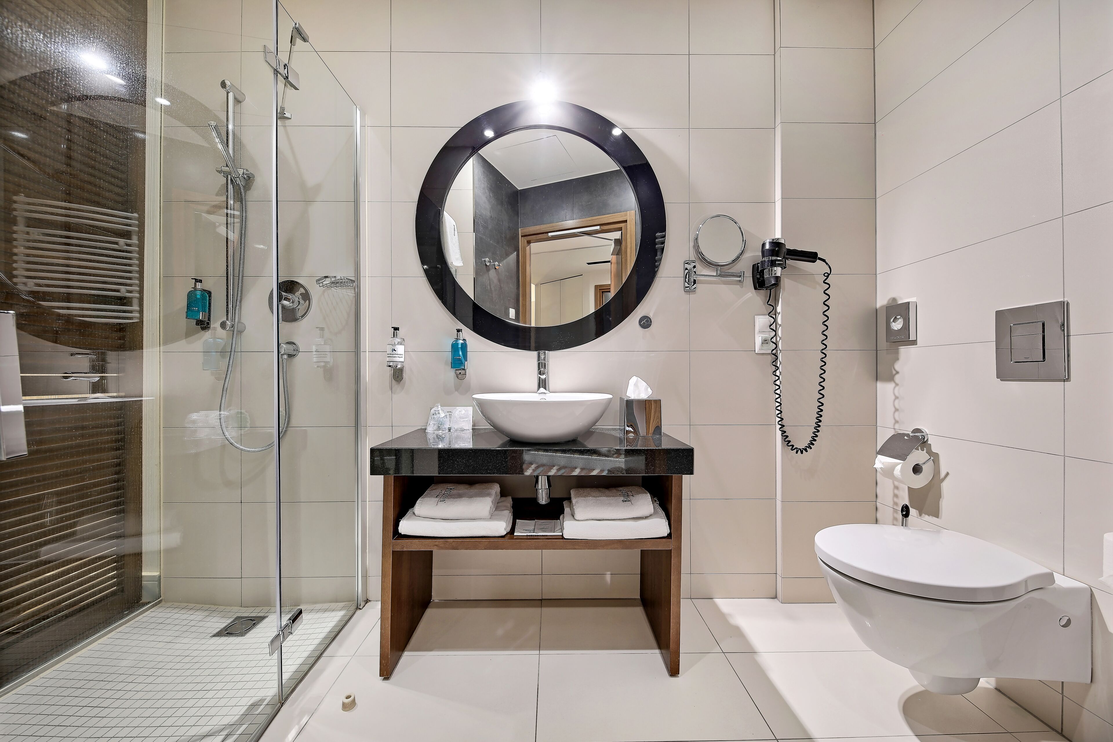 Une salle de bain moderne avec une douche à parois de verre, un miroir rond, un lavabo blanc et des toilettes.