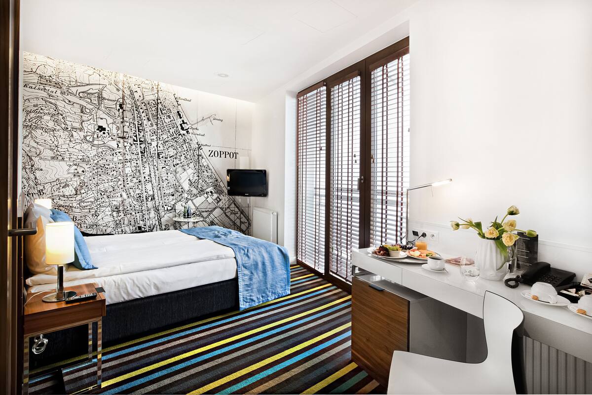 A modern hotel room with a large map on the wall, a bed with a blue blanket, a desk with a laptop, and a small table with a vase of flowers.