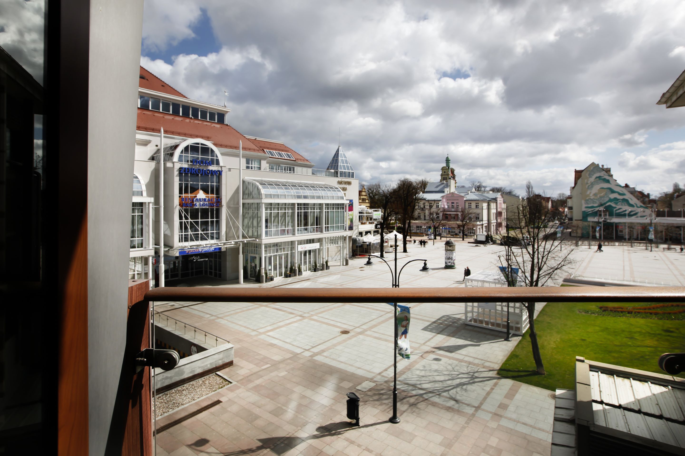 A spacious plaza with modern buildings, including a glass structure and a traditional building with a green roof.