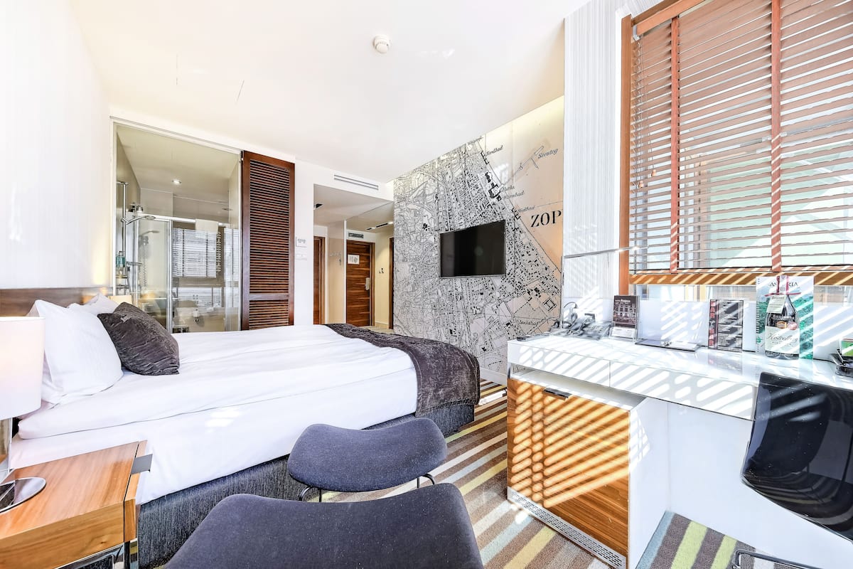 A modern hotel room with a large bed, a desk with a computer, a TV, and a map on the wall.