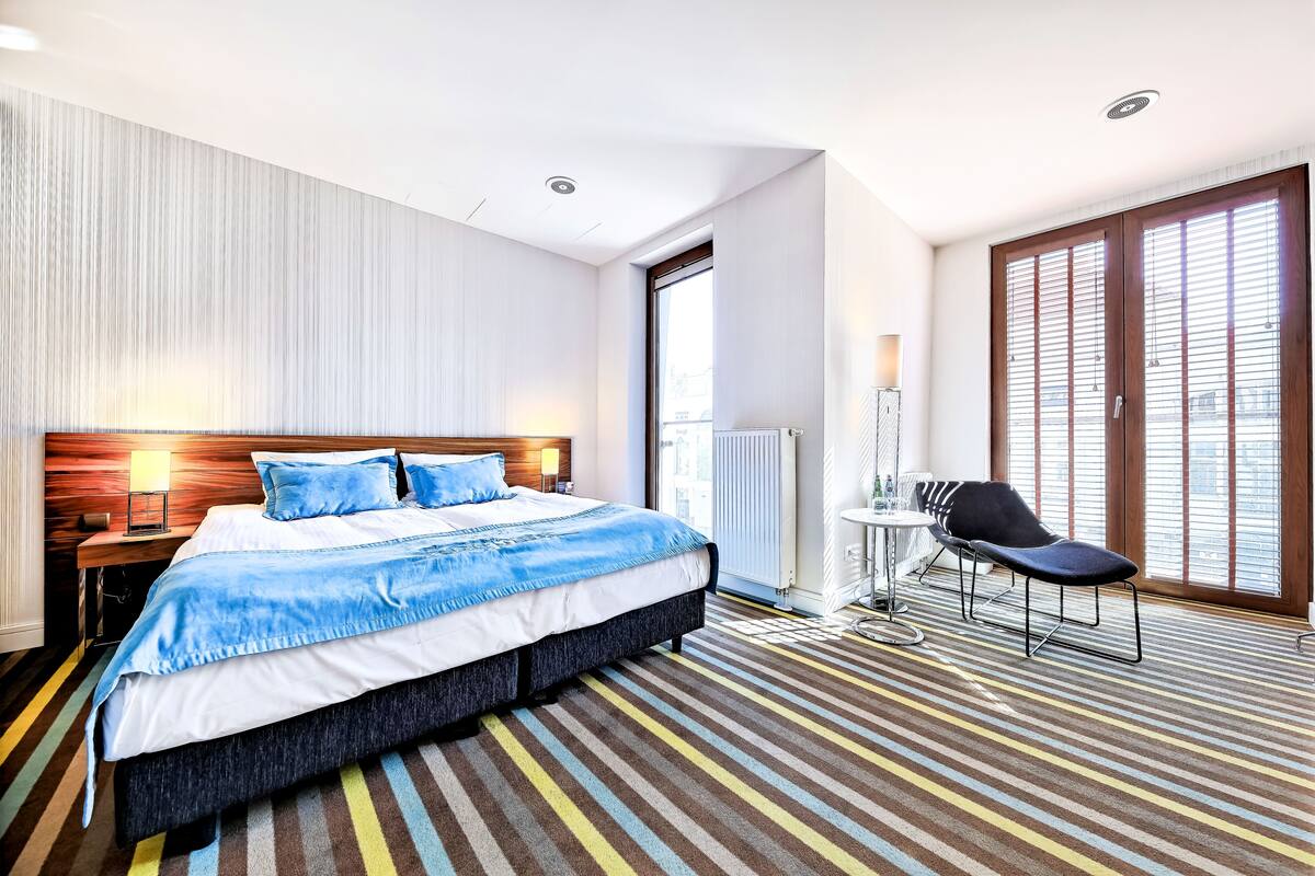 A modern hotel room with a large bed, a chair, a desk, and a view of the city through large windows.