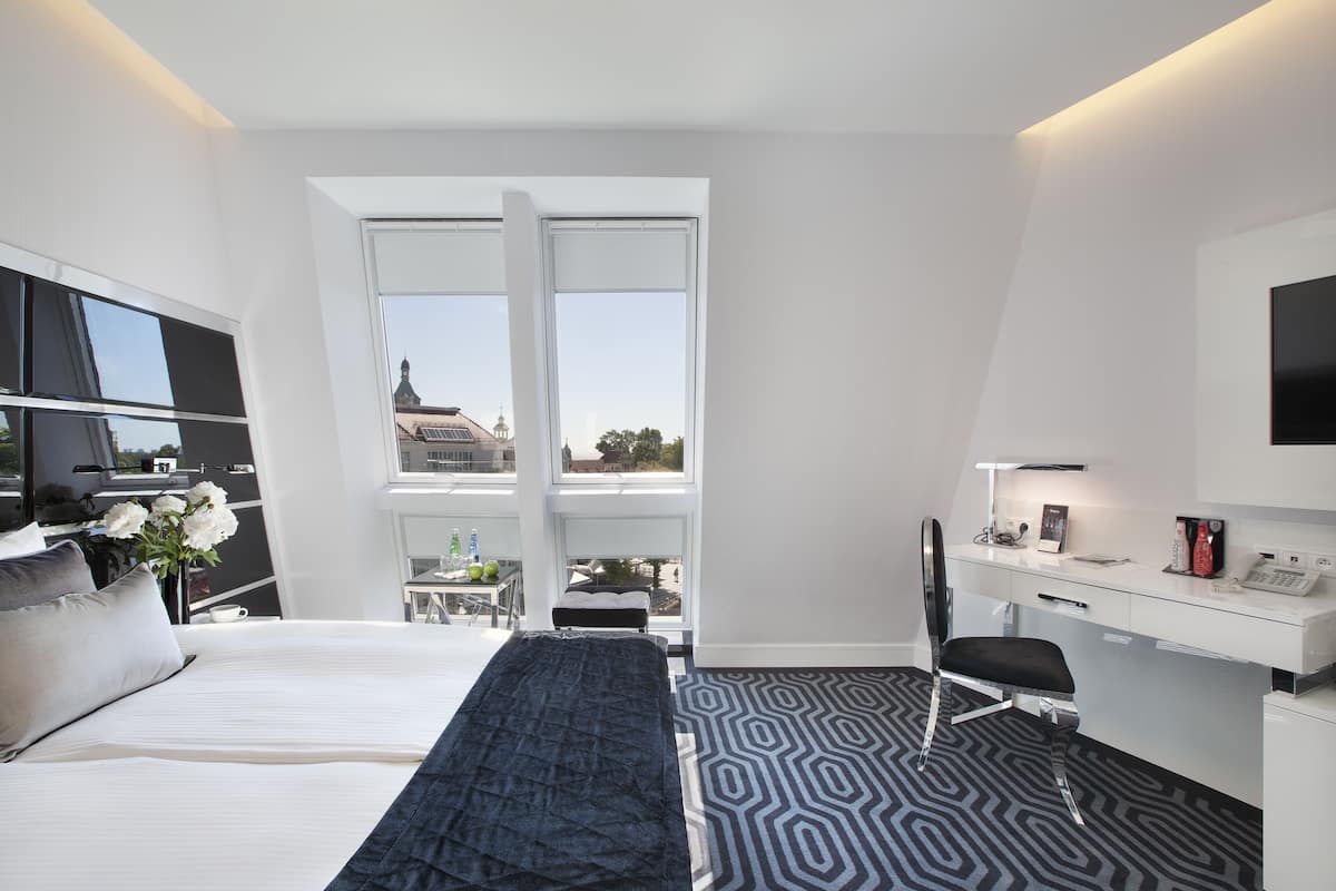 A modern hotel room with a large bed, a desk, and a view of the city through large windows.