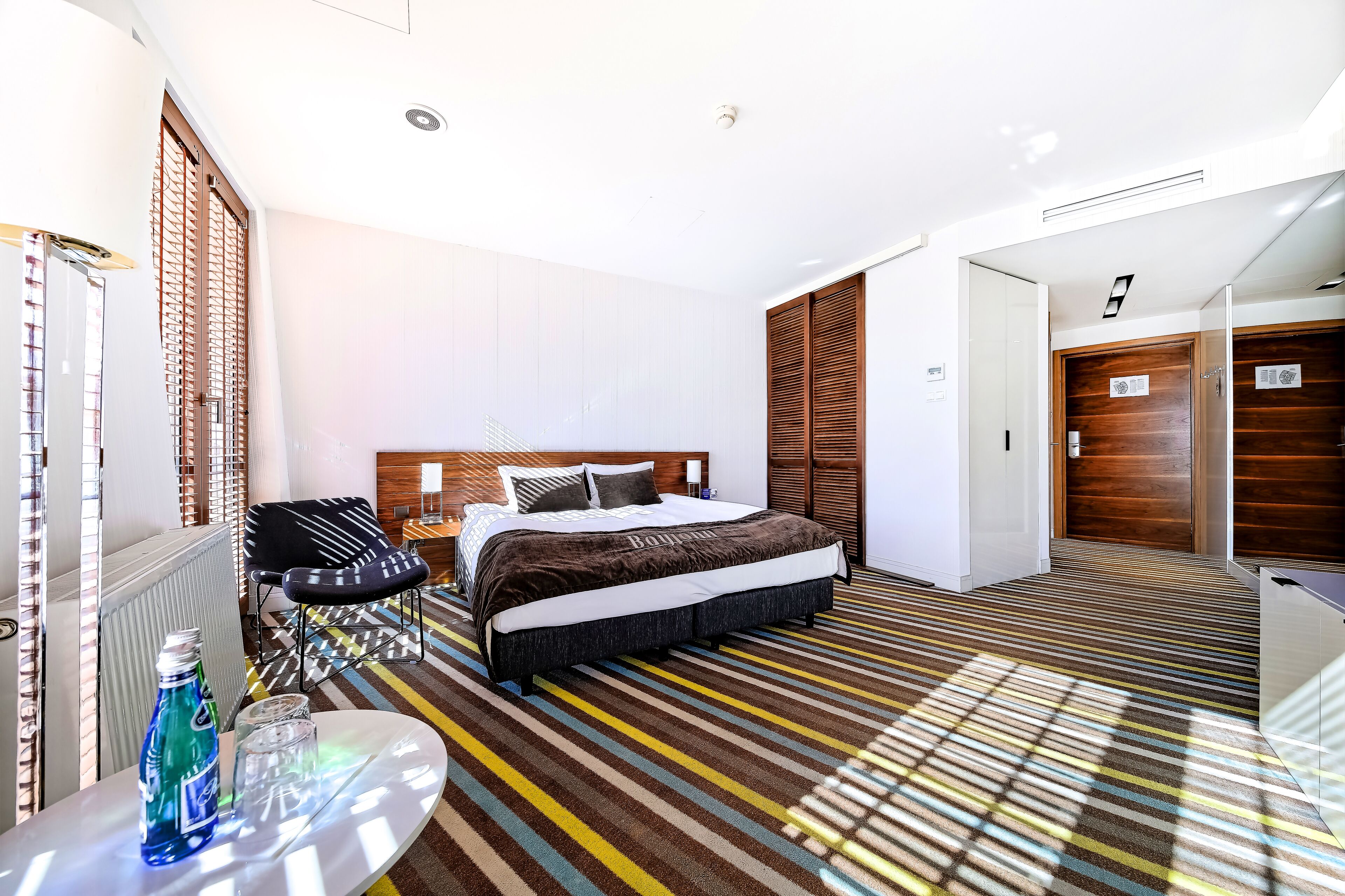 A hotel room with a bed, a chair, a table with a bottle, and a striped carpet.