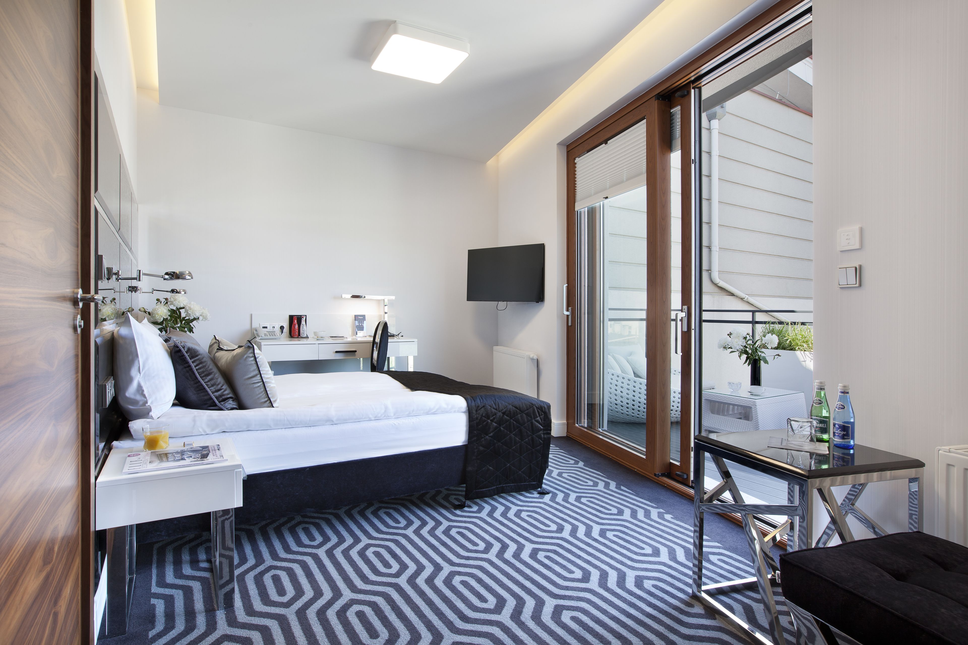 Una habitación de hotel moderna con una cama grande, un escritorio con una silla, un televisor montado en la pared y un ventanal corredero que da a un balcón.