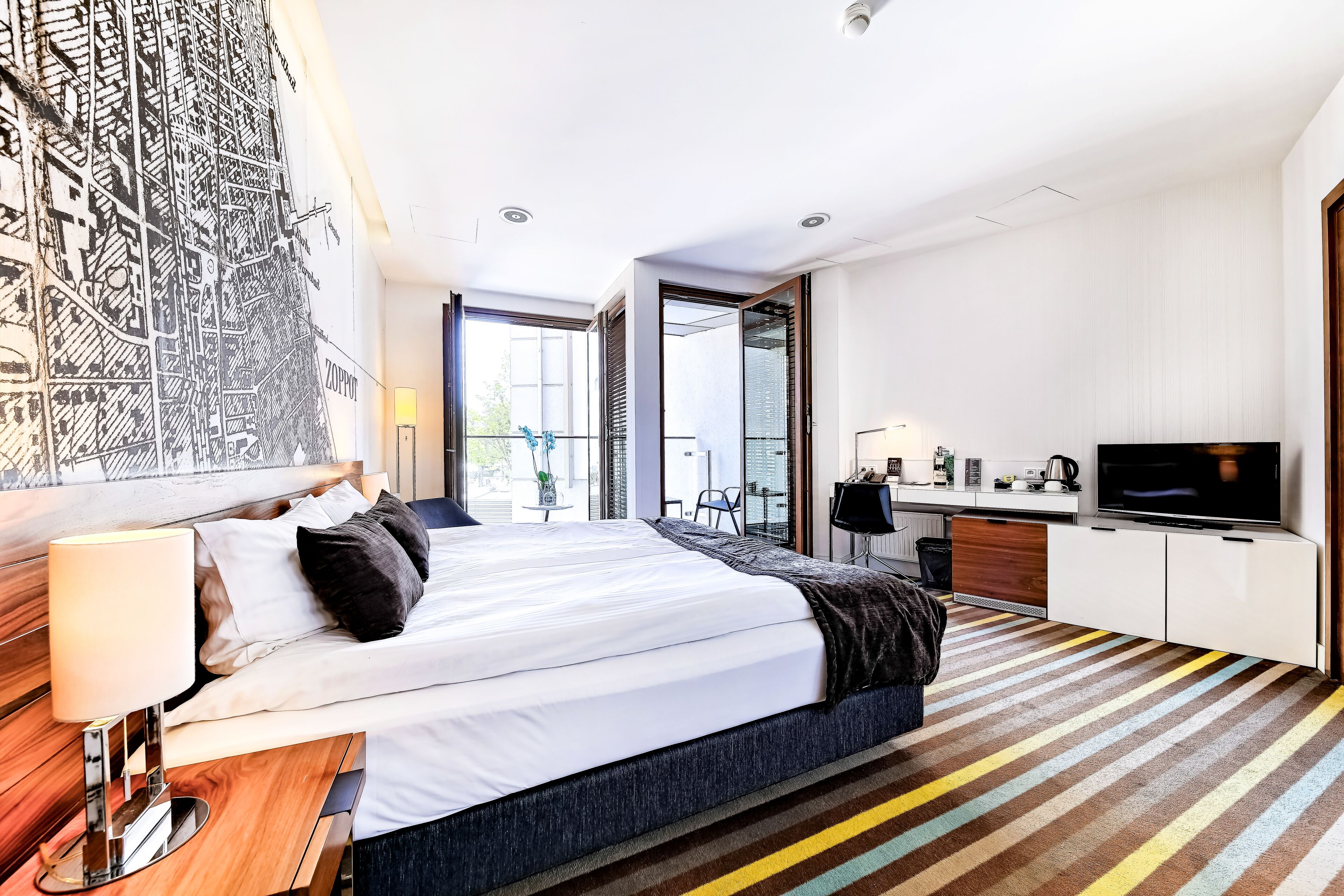 Une chambre d’hôtel moderne, dotée d’un grand lit, d’un bureau, d’une télévision et d’une fresque murale représentant un paysage urbain.