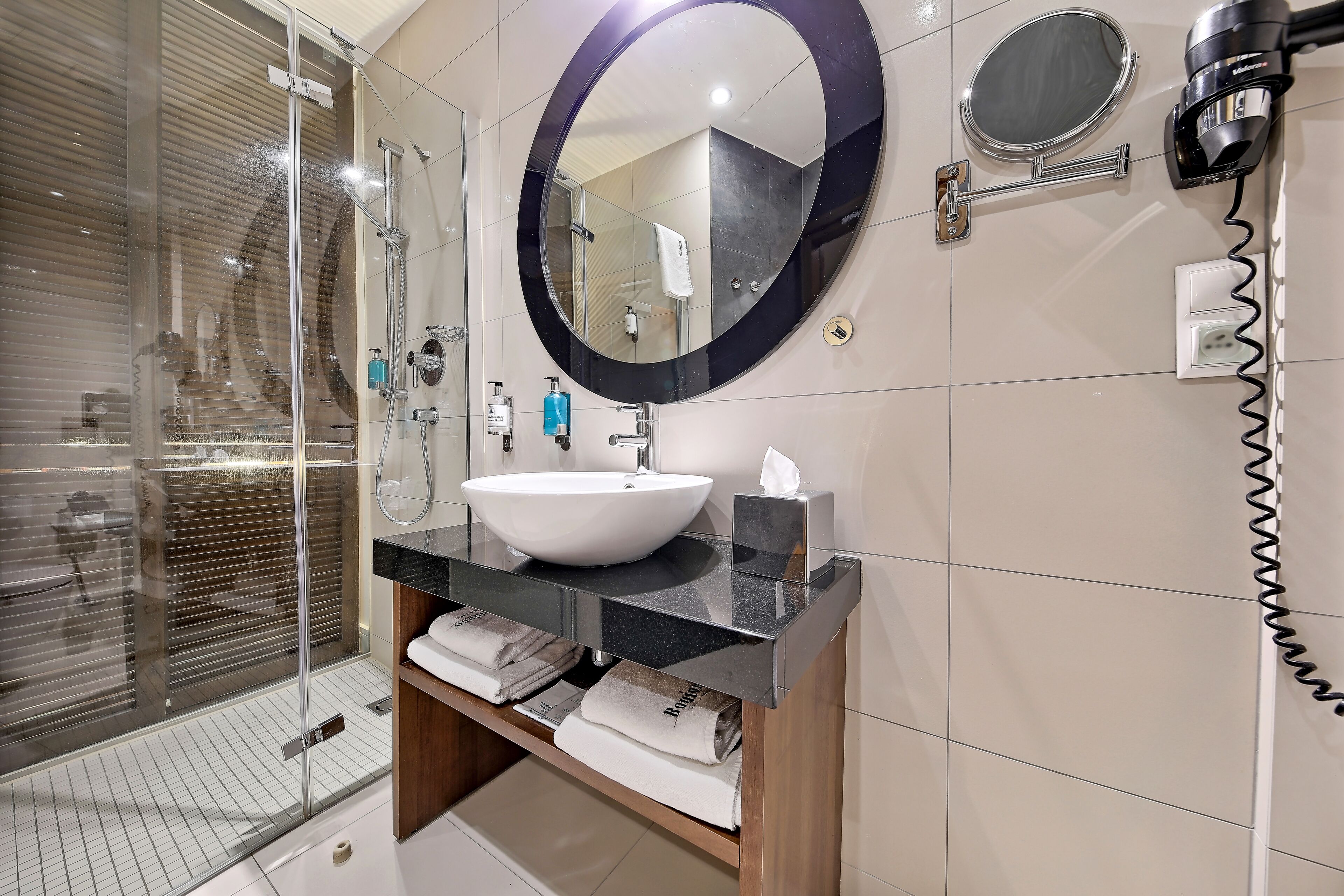A modern bathroom with a glass shower enclosure, a white sink, a round mirror, and a hairdryer mounted on the wall.