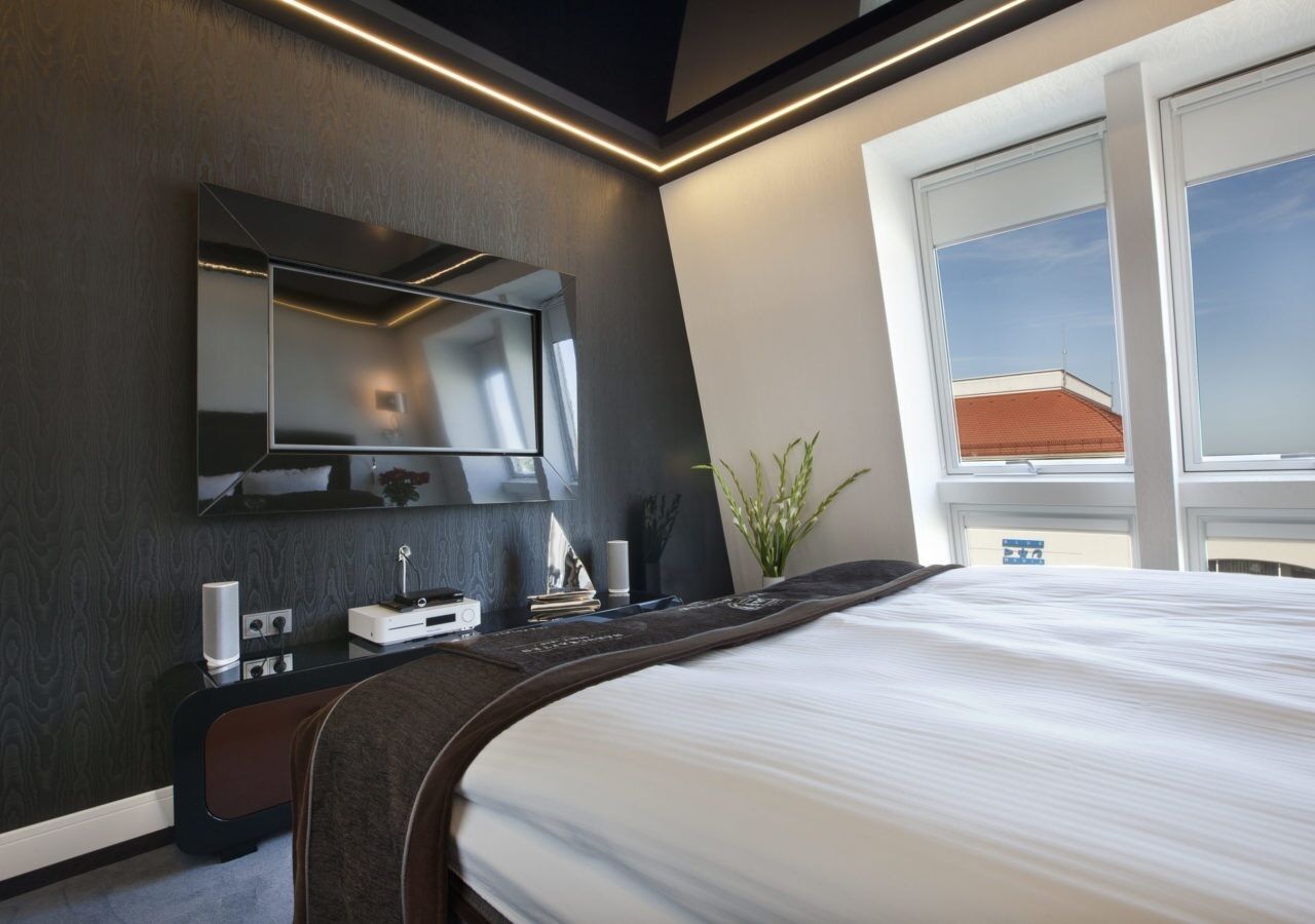Una habitación de hotel moderna con una cama grande, un televisor de pantalla plana en la pared, una mesita de noche con lámpara y una ventana con vistas al cielo.
