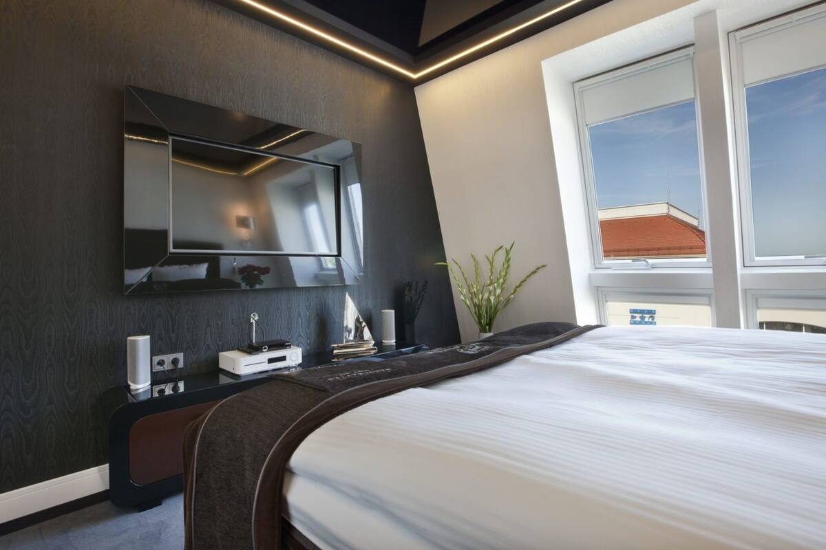 A modern hotel room with a large bed, a flat-screen TV mounted on the wall, a bedside table with a lamp, and a window with a view of the sky.