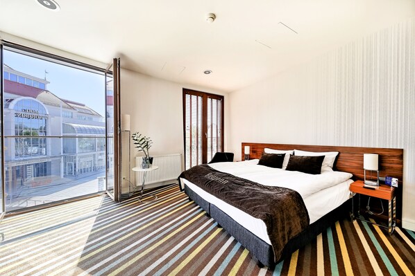 A hotel room with a large bed, a bedside table with a lamp, a view of a building outside, and a striped carpet.