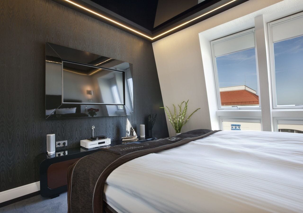 Una habitación de hotel moderna con una cama grande, un televisor de pantalla plana en la pared, una mesita de noche con lámpara y una ventana con vistas al cielo.