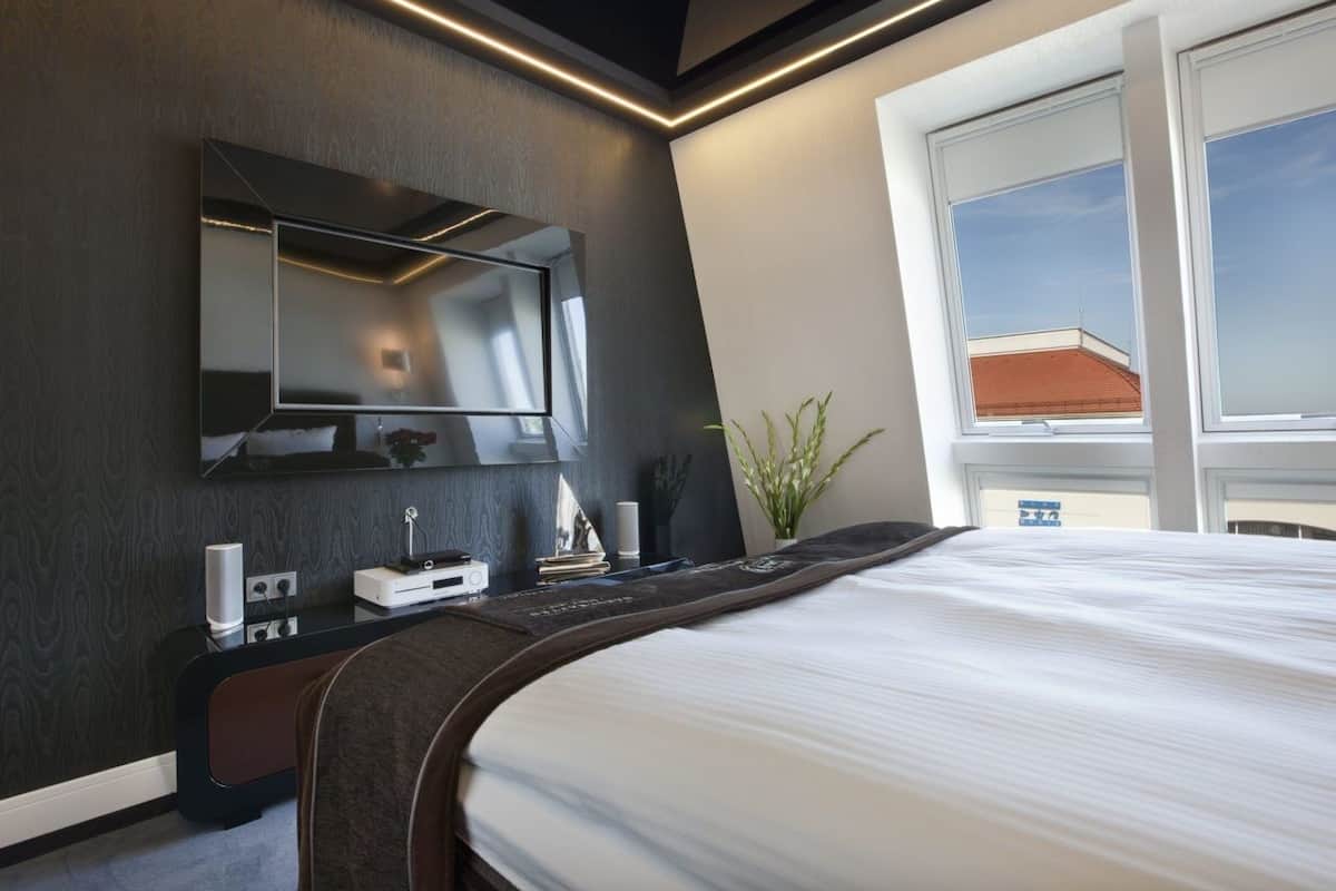 A modern hotel room with a large bed, a flat-screen TV mounted on the wall, a bedside table with a lamp, and a window with a view of the sky.