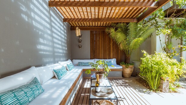 Un espacio de estar al aire libre iluminado por el sol, con una pérgola de madera, un sofá con cojines, una mesa con una botella y vasos, y diversas plantas en macetas.