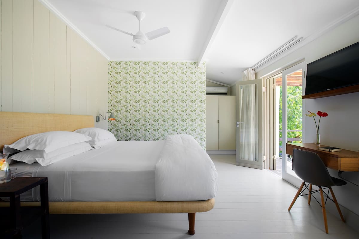 A bedroom with a bed, a desk, a chair, a TV, and a window with a view of greenery.