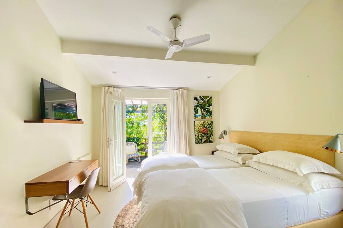 A bedroom with two beds, a desk, a chair, a ceiling fan, and a view of greenery outside.