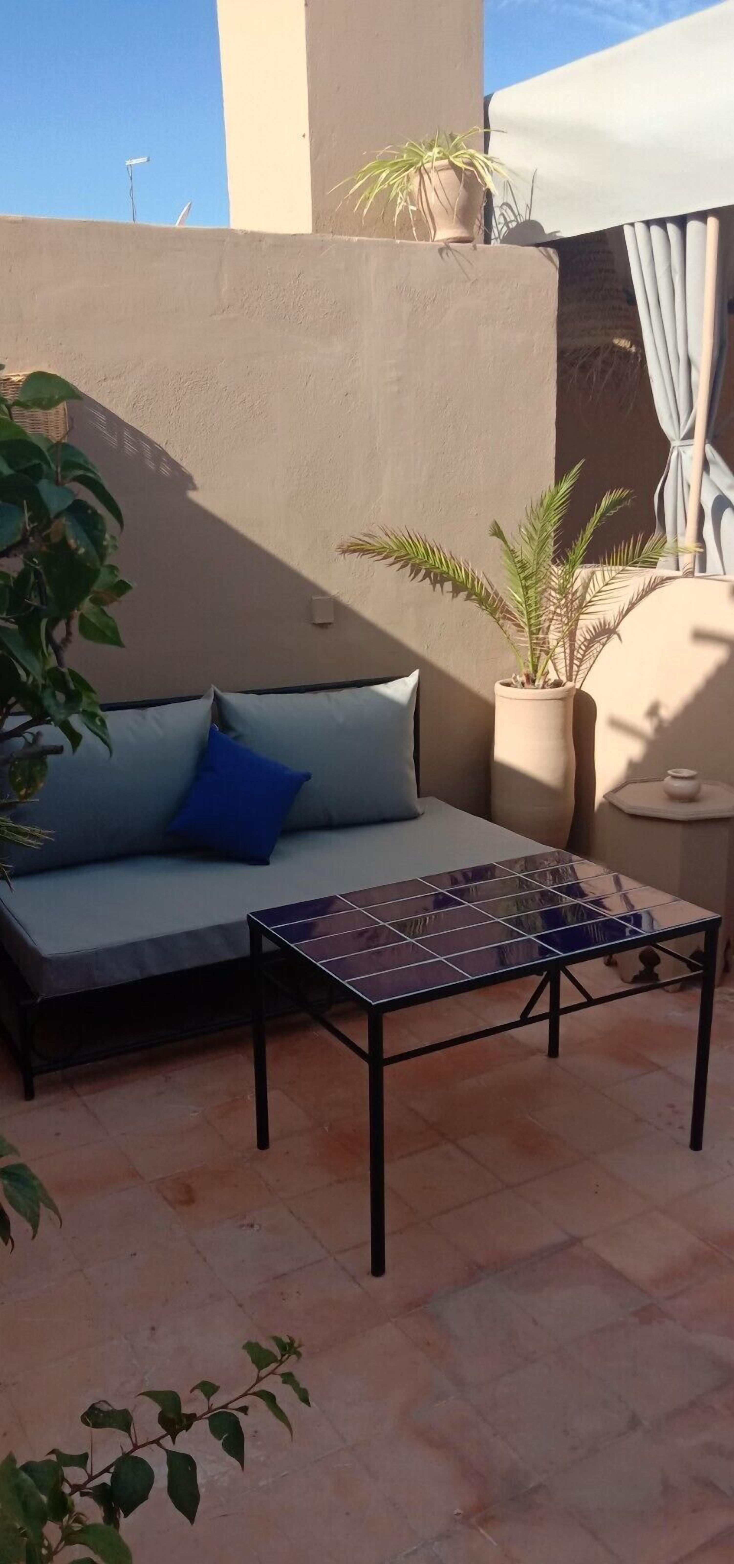 Ein sonnendurchfluteter Außenbereich mit einer Sitzecke, einem Sofa und einem kleinen Tisch, umgeben von Topfpflanzen und einem gefliesten Boden.