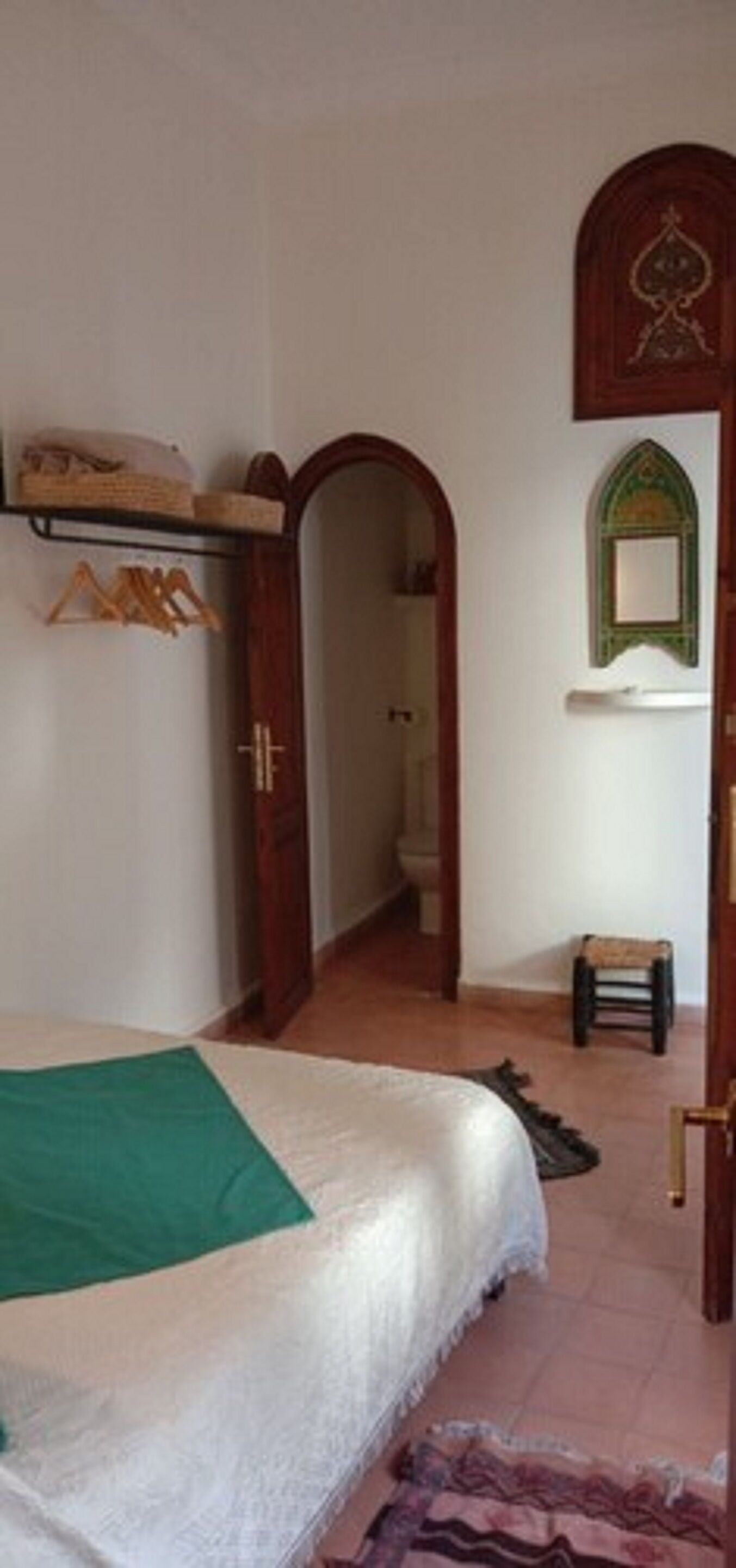A bedroom with a bed, a wooden wardrobe, a green throw, a small stool, and a mirror on the wall.