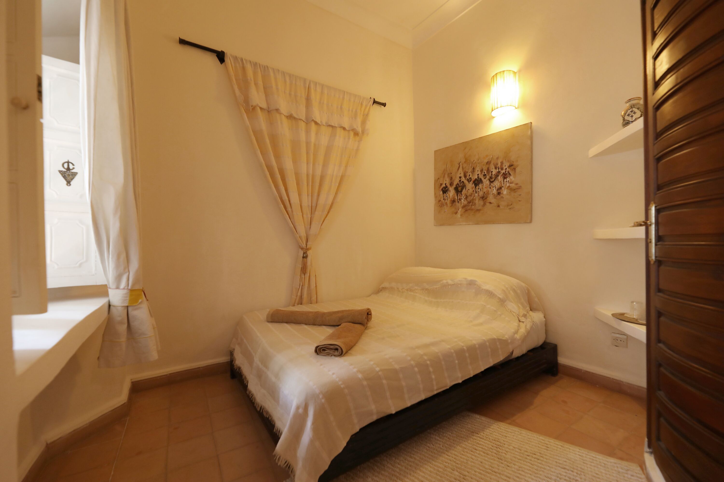 Une chambre à coucher comprenant un lit, une table de chevet, une lampe fixée au mur, un tableau et une fenêtre avec des rideaux.