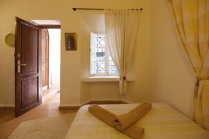 A bedroom with a bed, a window with curtains, a wooden door, and a small window with decorative ironwork.