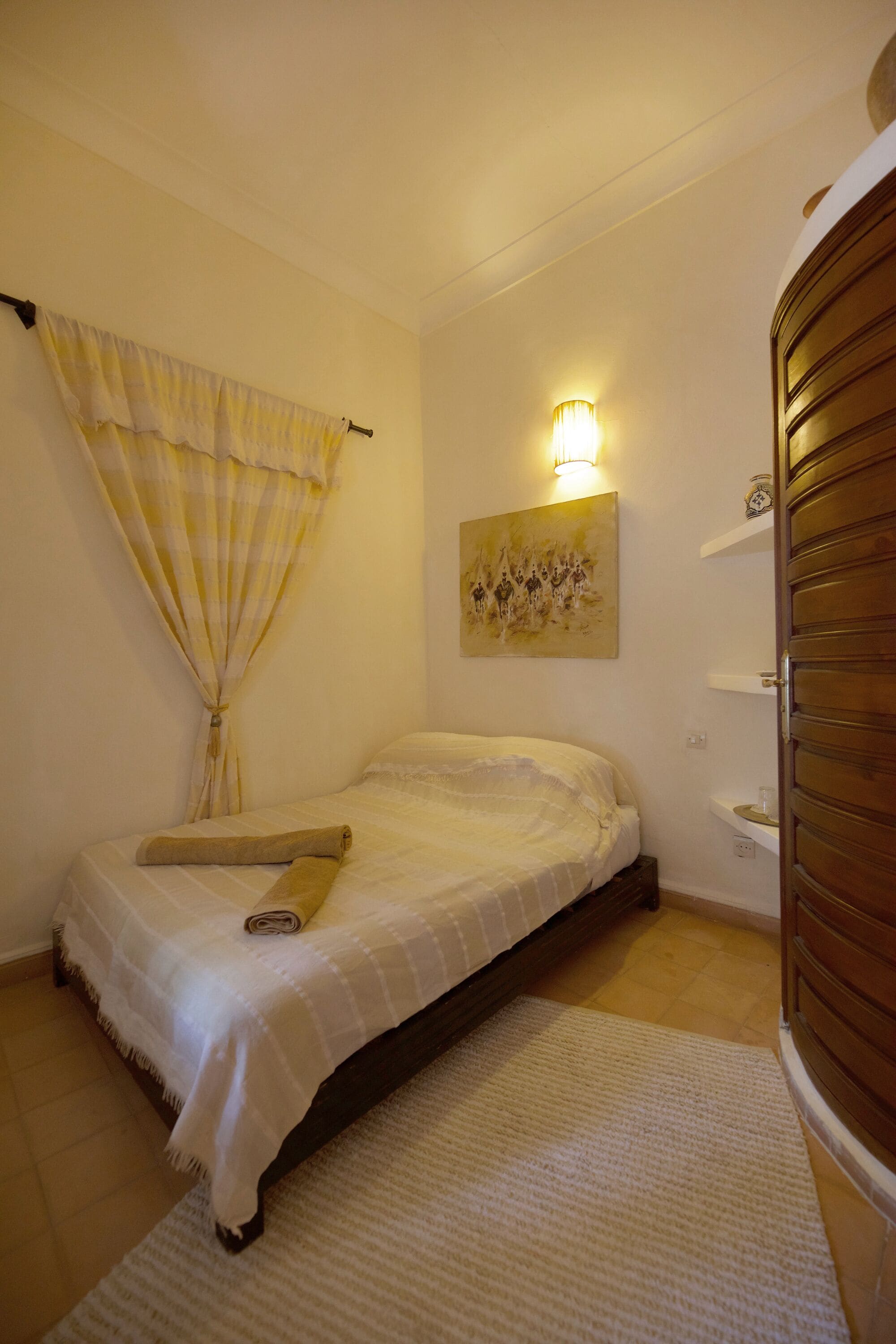 Une chambre à coucher avec un lit, une fenêtre avec des rideaux, une lampe fixée au mur, un tableau et une armoire en bois.