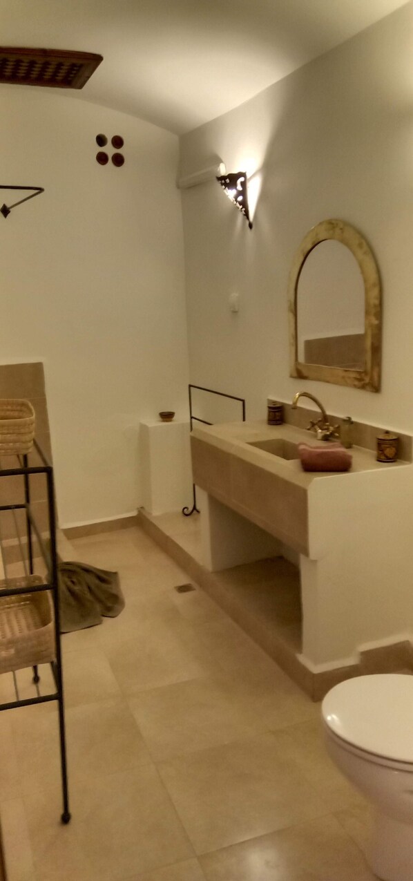 Un baño con lavamanos, espejo e inodoro. Hay un perchero y un estante pequeño con artículos de aseo.