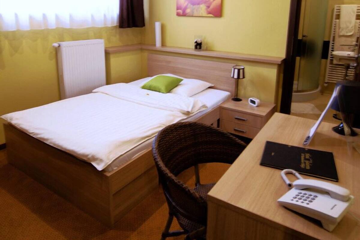 A hotel room with a bed, a desk with a telephone, a chair, a bedside table with a lamp, and a small shelf with a decorative item.